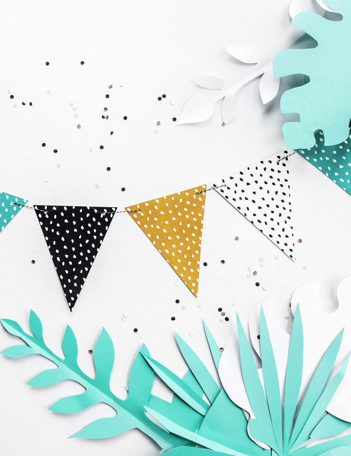Mise en situation avec feuilles tropicales de la guirlande de fanions à pois blanc pour fête multicolore en carton